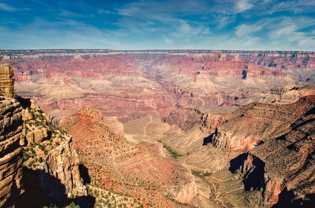 A breathtaking view of the Grand Canyon in Arizona during summer vacation, showcasing the vast, colorful landscape with deep gorges and layered rock formations under a clear blue sky. Families can be seen enjoying the scenery and taking photographs at a popular viewpoint, emphasizing the destination's appeal as a family-friendly spot for summer explorations.