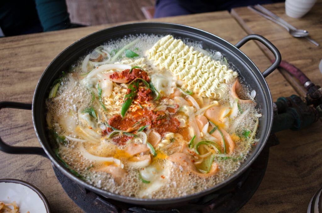 Combining ramen noodles and seasoning in a pot, stirring to coat evenly. Steam rises from the pot, showcasing a flavorful preparation process.