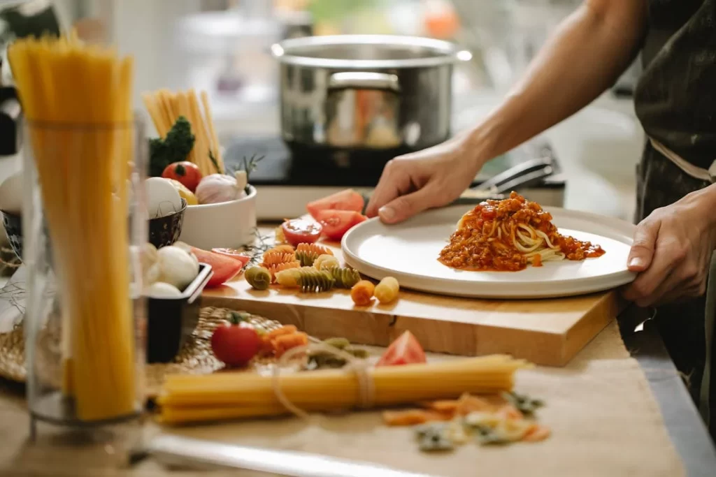 "Photo of a home kitchen setting where fresh homemade spaghetti is being prepared. Ingredients like flour, eggs, and olive oil are visible on the countertop. In the center, a person rolls out pasta dough with a wooden rolling pin. The image captures the process of cutting the dough into thin spaghetti strands, emphasizing the hands-on method of crafting traditional spaghetti."