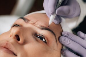 "Close-up of a professional beautician applying wax to a client's eyebrows in a clean, well-lit salon setting, demonstrating the technique of brow waxing. The image captures the precise application of wax strips under the guidance of an expert, highlighting the care and attention required for successful brow waxing."