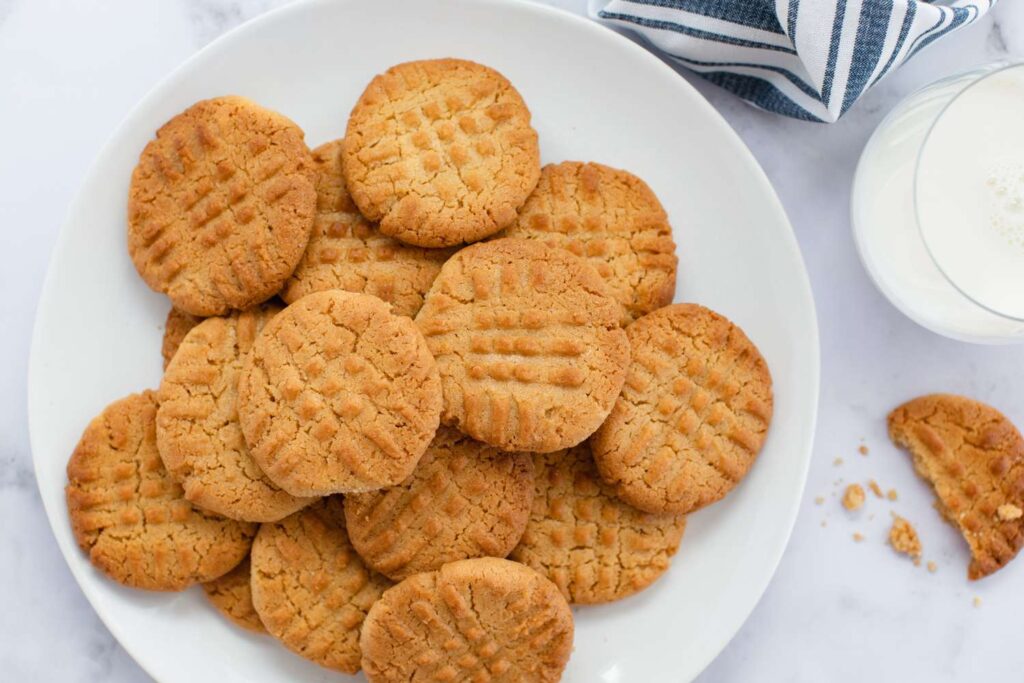 Classic Peanut butter cookies, golden brown with a soft, crumbly texture, neatly arranged on a plate. Each cookie features the signature crisscross pattern on top, emphasizing their homemade charm. The rich, nutty aroma of peanut butter wafts from the cookies, highlighting their key ingredient. These Peanut butter cookies are a perfect blend of sweetness and savory peanut flavor, ideal for a nostalgic treat or a simple dessert.