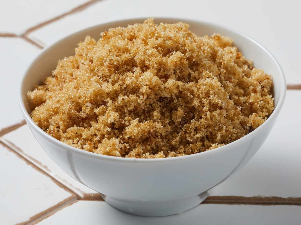 Close-up of a hand gently sprinkling rich brown sugar into a mixing bowl filled with banana bread batter, giving the bread extra moisture and a hint of caramel sweetness. The brown sugar is being added to create the perfect balance of flavor and texture for a truly moist banana bread