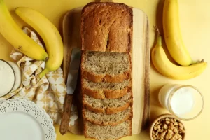 A close-up view of a slice of moist banana bread with a golden-brown crust and a soft, fluffy interior, showcasing its rich texture. The banana bread is studded with visible banana chunks, giving it a deliciously moist and tender appearance, perfect for a comforting snack or breakfast.