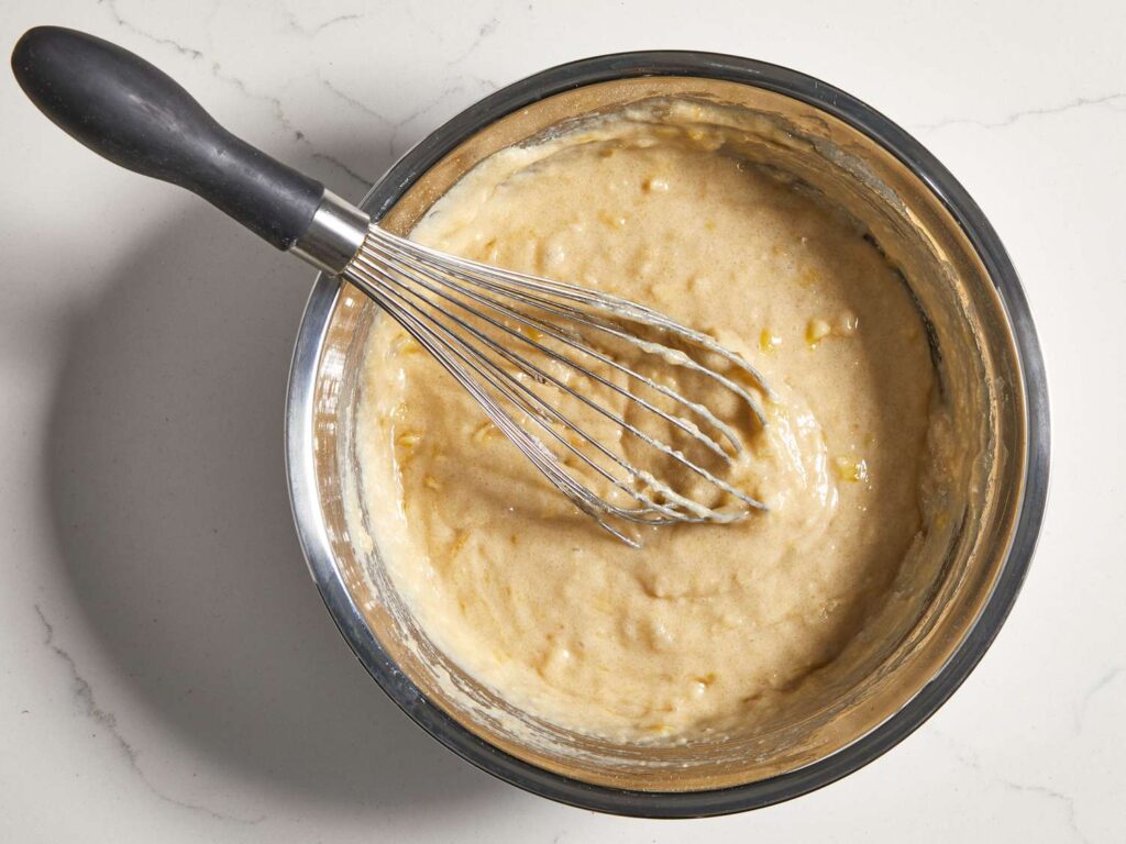 Close-up of moist banana bread batter being gently stirred with a spatula in a mixing bowl. The smooth texture of the batter looks creamy and thick, with small lumps indicating that it's not overmixed. The key to this moist banana bread is to fold the ingredients just enough to combine, ensuring a fluffy, tender loaf once baked. The bowl and utensils create a cozy baking atmosphere, emphasizing the importance of not overmixing for perfect moisture and texture.