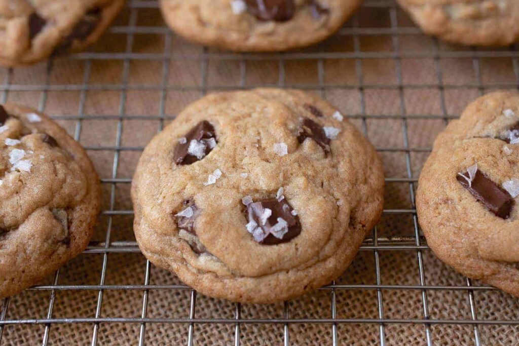 A close-up of freshly baked chocolate chip cookies sprinkled with flaky sea salt, showcasing their golden-brown edges and melty chocolate chips, resting on a rustic wooden surface.