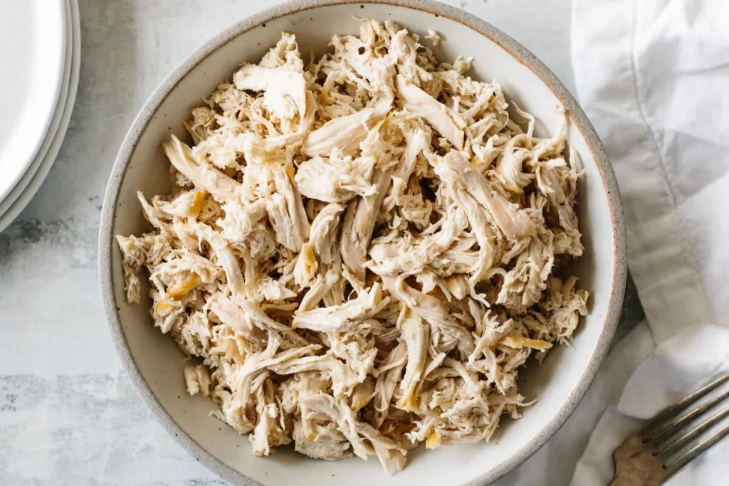 A close-up image of a fork shredding tender, cooked chicken breast, showcasing the process of preparing ingredients for a delicious Chicken Salad Sandwich. The shredded chicken is placed in a mixing bowl, surrounded by fresh vegetables and mayonnaise, highlighting the essential steps in making the perfect Chicken Salad Sandwich.