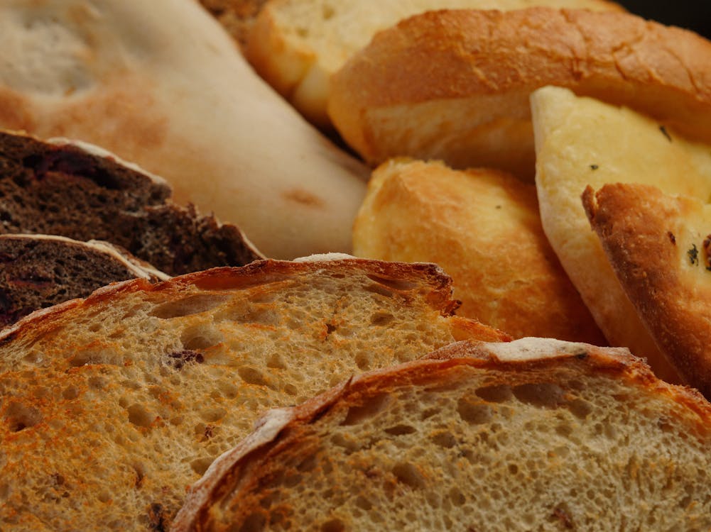 A selection of thick-cut bread varieties ideal for making French Toast, including brioche, challah, and French baguette. The bread is arranged on a wooden cutting board with a whisk, eggs, and milk in the background, ready to be transformed into rich and flavorful French Toast.