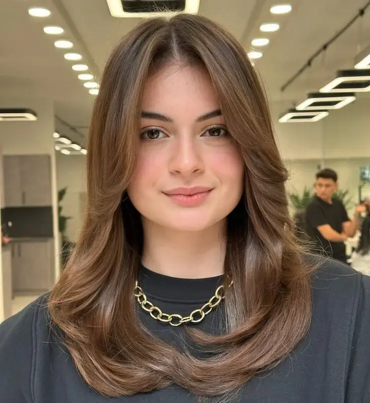 Image of a woman with long, flowing hair styled with curtain bangs and soft layers. The haircut features voluminous curtain bangs that frame her face, blending seamlessly into the long layers. This trendy and versatile hairstyle enhances facial features and adds movement to the hair. Perfect for those looking for a low-maintenance yet stylish look, this 'Curtain Bangs with Long Layers' cut is one of the popular Haircut Styles for Women.